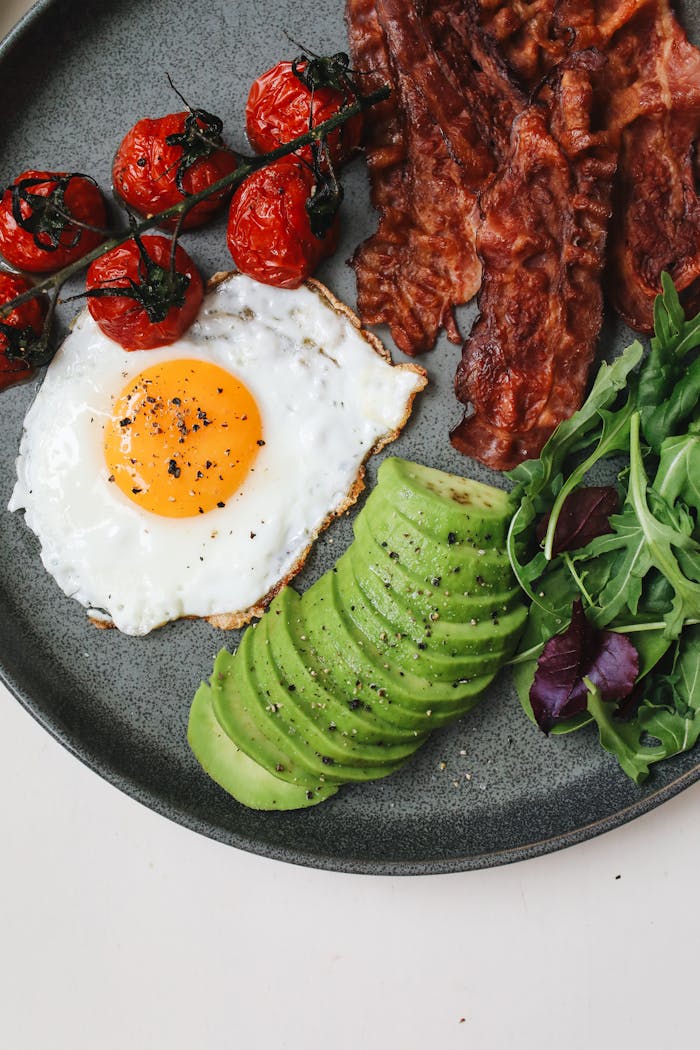 Delicious breakfast of crispy bacon, sunny-side egg, avocado slices, tomatoes, and greens.